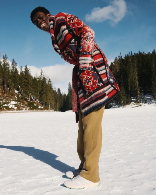 Adult male in a patterned knit cardigan and tan trousers bends forward smiling on a snowy plain under a blue sky with a forest visible.