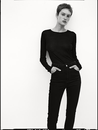 Model with short hair wearing a black long-sleeve top and dark trousers against a white seamless background.