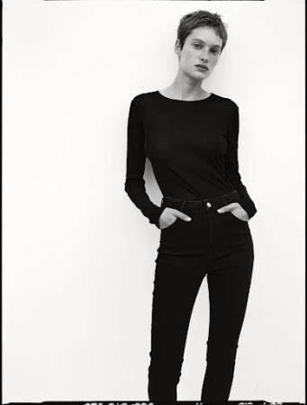Model with short hair wearing a black long-sleeve top and dark trousers against a white seamless background.