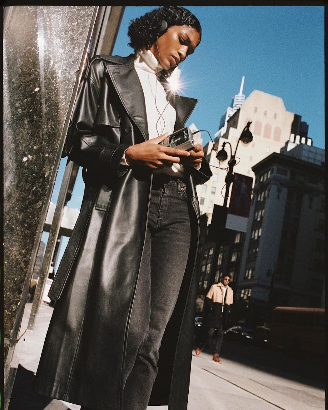 A woman in a black trench coat holds a vintage Walkman on an urban street with buildings and blue sky in the background on a sunny day.