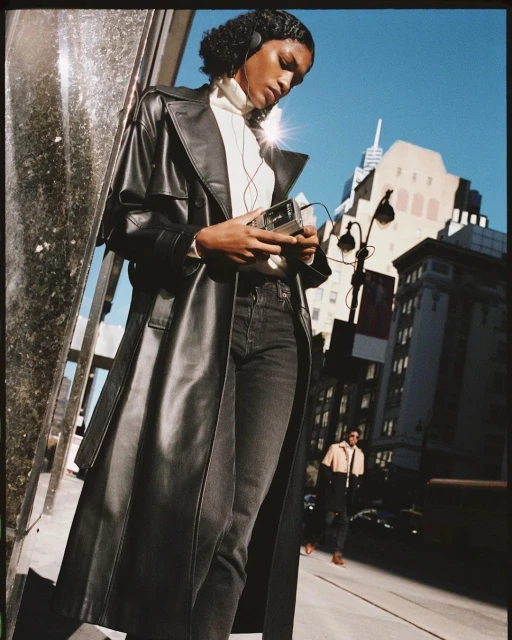 A woman in a black trench coat holds a vintage Walkman on an urban street with buildings and blue sky in the background on a sunny day.
