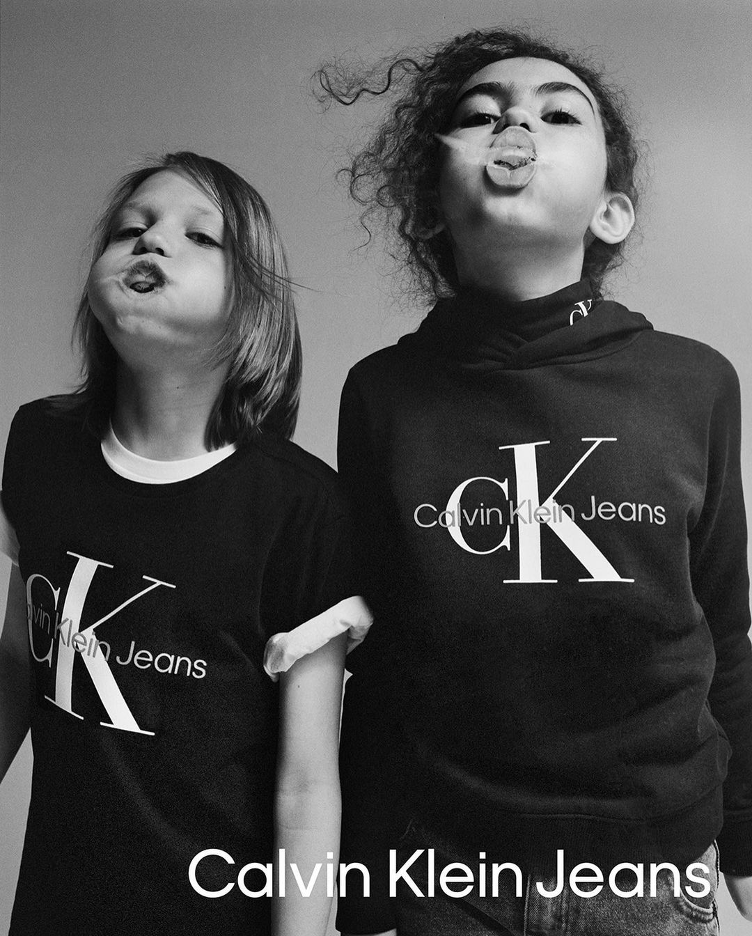 Two children pose indoors wearing Calvin Klein Jeans apparel, one in a t-shirt and one in a hoodie, sticking out their tongues.