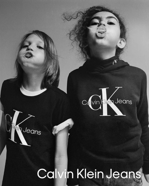 Two children pose indoors wearing Calvin Klein Jeans apparel, one in a t-shirt and one in a hoodie, sticking out their tongues.