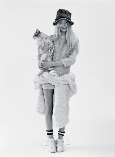 Young girl in hat poses with tabby cat in bright studio, wearing sweatshirt, socks, sneakers, and white jacket around her waist.