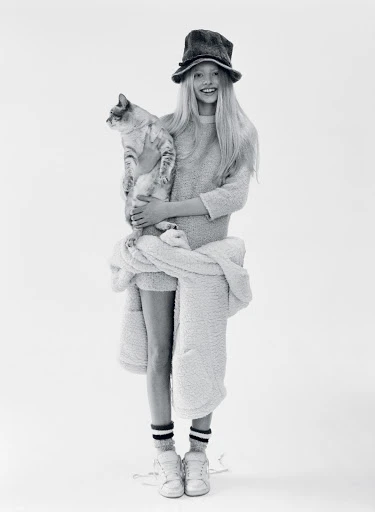 Young girl in hat poses with tabby cat in bright studio, wearing sweatshirt, socks, sneakers, and white jacket around her waist.