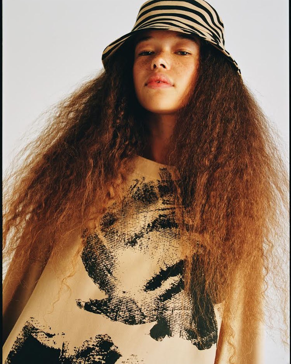 Young adult female poses in a tan dress and zebra-striped bucket hat against a white backdrop in a studio-lit fashion shot.