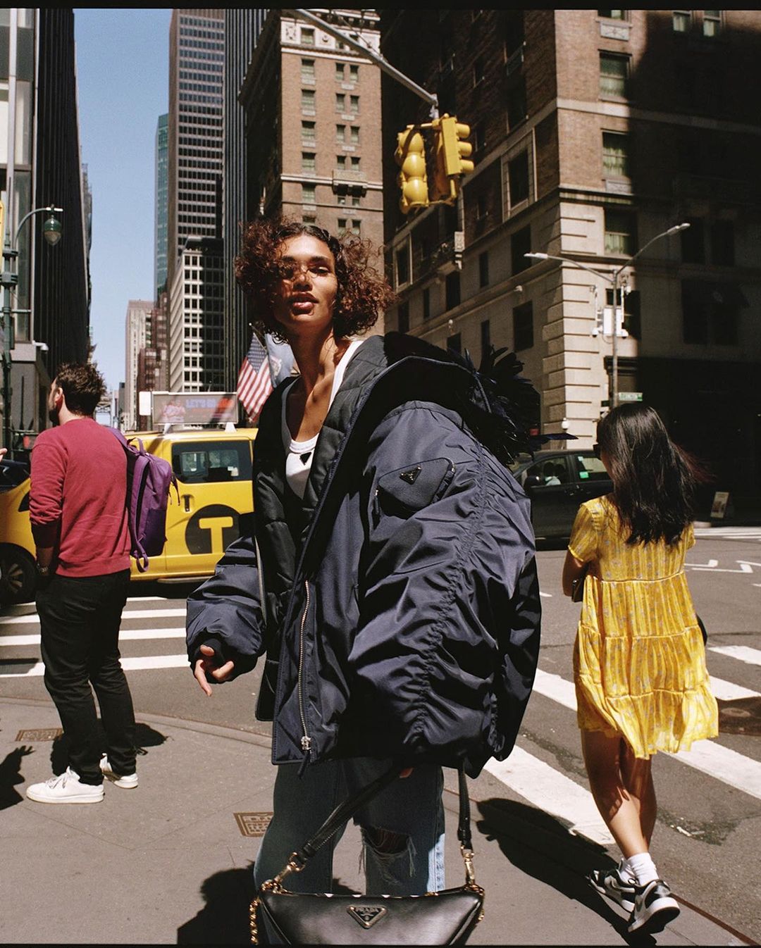 A woman in a navy bomber jacket and jeans crosses a New York City street. Taxis and pedestrians fill the background on a sunny day.