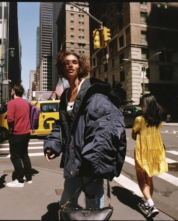 A woman in a navy bomber jacket and jeans crosses a New York City street. Taxis and pedestrians fill the background on a sunny day.