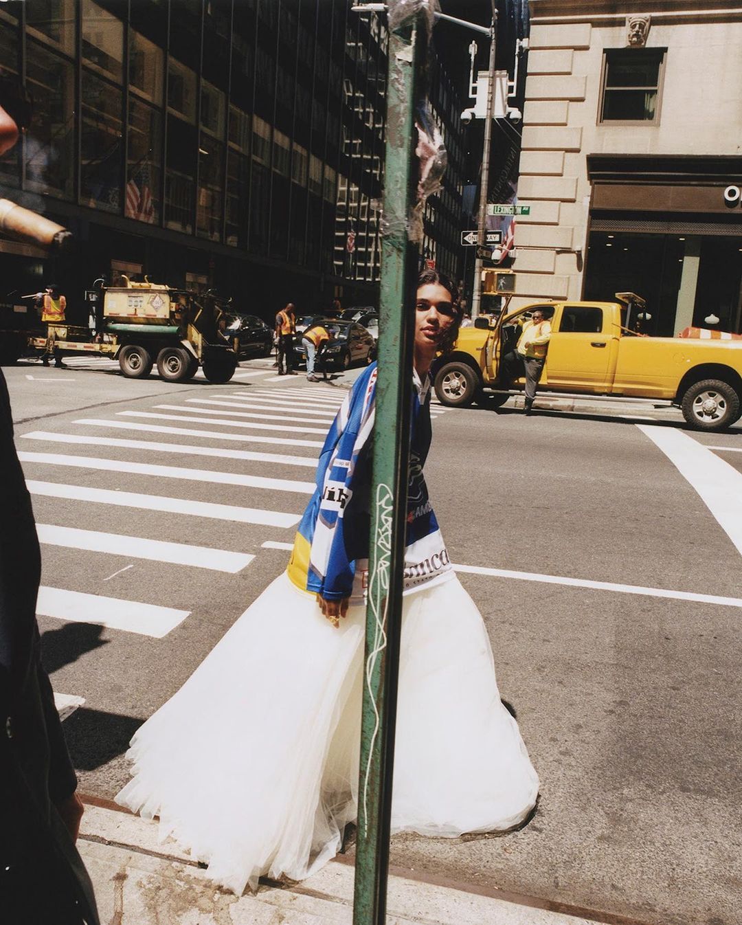 Woman in a dress walks on a city street crosswalk, wearing a blue and yellow jacket with city buildings and vehicles in the background.