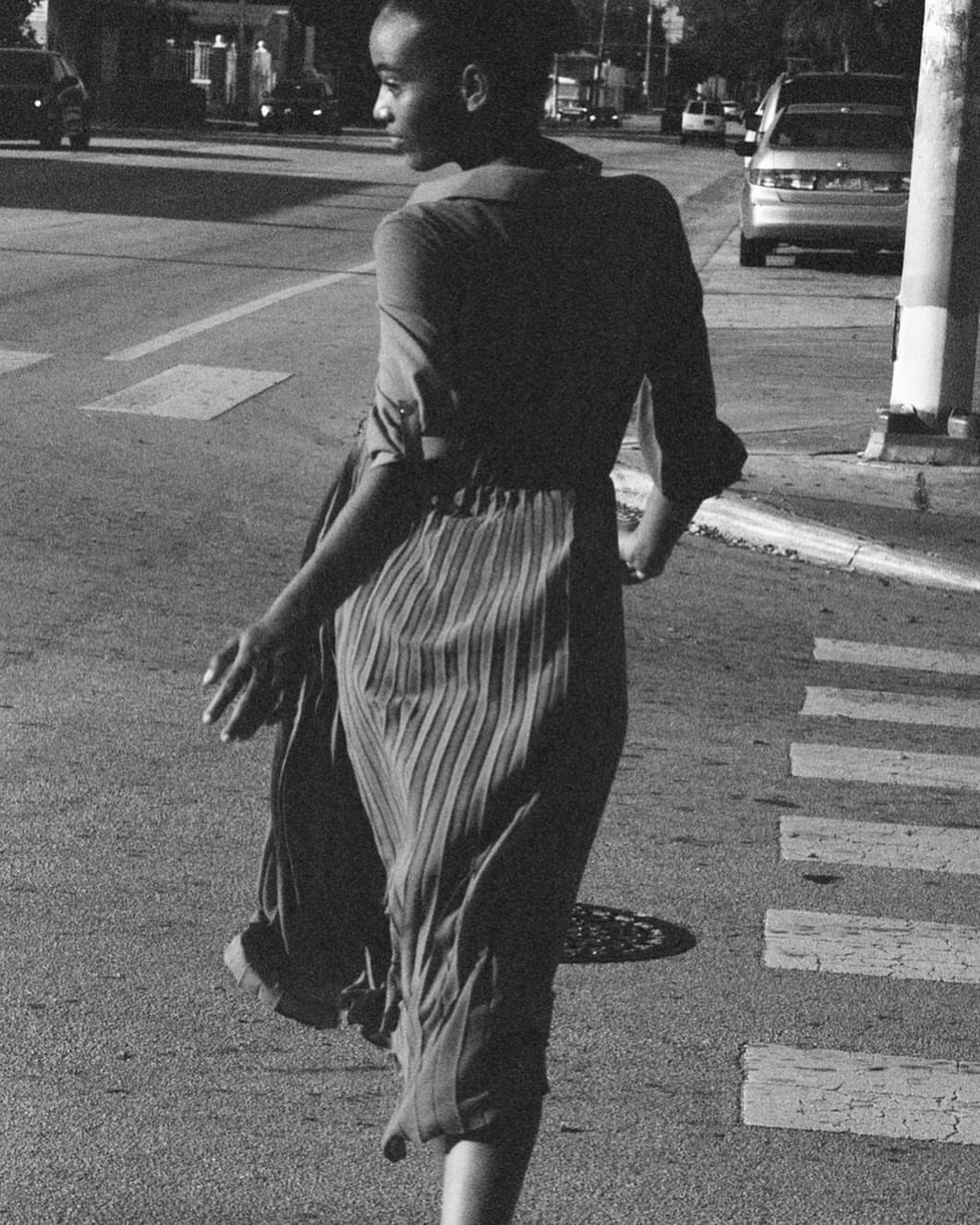 A person is seen from behind, crossing a crosswalk in a black-and-white outdoor photograph on a city street with cars in the background.