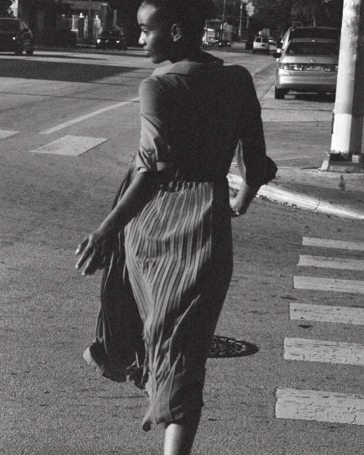 A person is seen from behind, crossing a crosswalk in a black-and-white outdoor photograph on a city street with cars in the background.