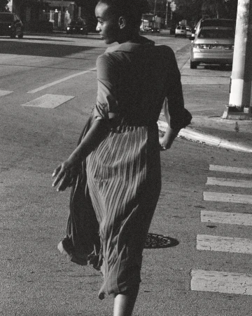 A person is seen from behind, crossing a crosswalk in a black-and-white outdoor photograph on a city street with cars in the background.