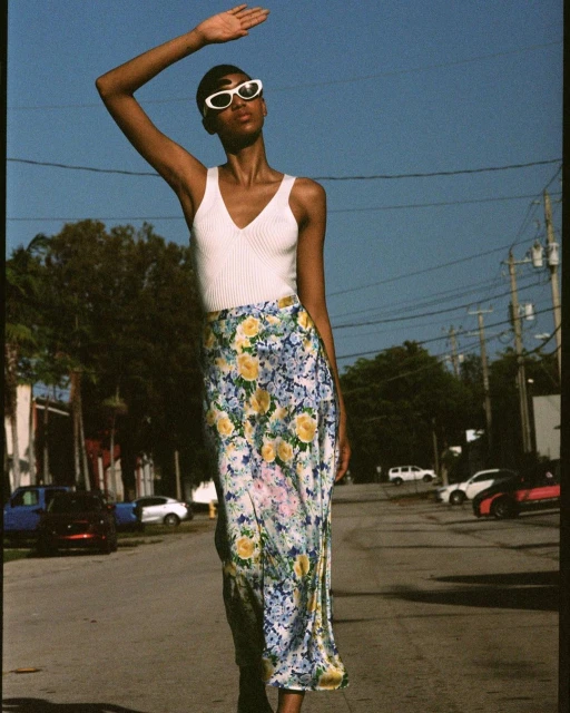 Woman in summer fashion stands on street in floral skirt and white top, blocking the sun with her hand under a blue sky. 