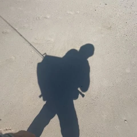 Shadow of an adult person with a backpack and fishing rod on a light-colored sandy beach in the daylight.