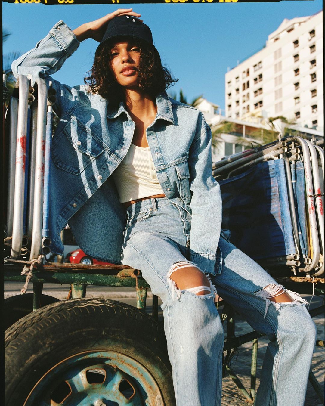 A young woman sits on a metal cart wearing a denim jacket and ripped jeans, with a building and palm trees in the background under a blue sky.