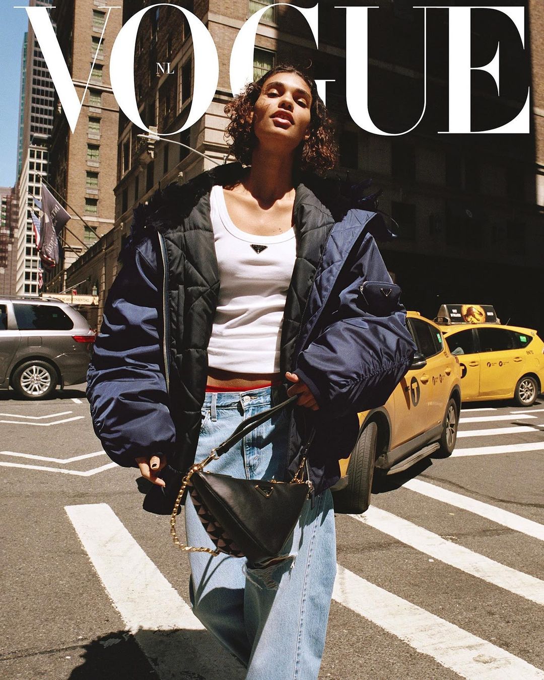 Fashion shot featuring a woman in a white top and blue jeans crossing a New York City street with a handbag, skyscrapers in background.