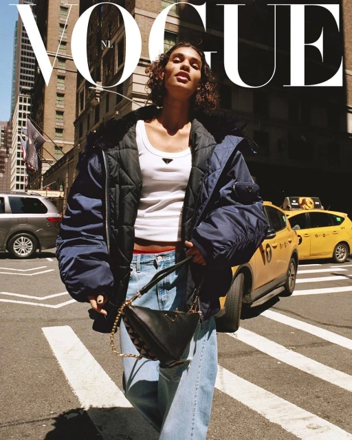 Fashion shot featuring a woman in a white top and blue jeans crossing a New York City street with a handbag, skyscrapers in background.