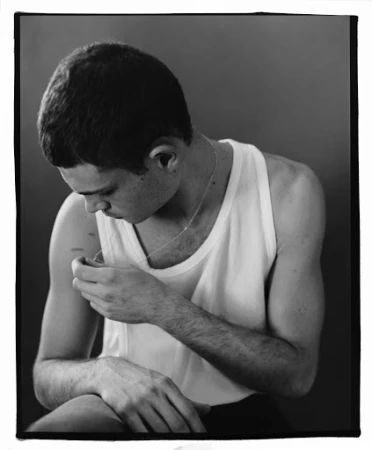 Monochrome portrait of a young man wearing a white tank top looking down while adjusting a thin necklace.