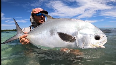 Fisherman holding a large permit fish in shallow, clear water with a blue sky. He wears sunglasses and a fishing hat. Clear water surrounds him.