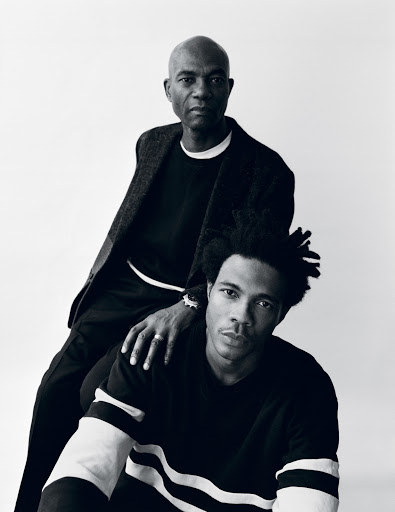 Portrait of an adult father with hand on shoulder of his adult son, both gazing forward in a monochrome photo studio setting.