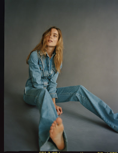 An adult woman with long blonde hair is sitting on a gray surface wearing a light blue denim jumpsuit, and one bare foot is visible.
