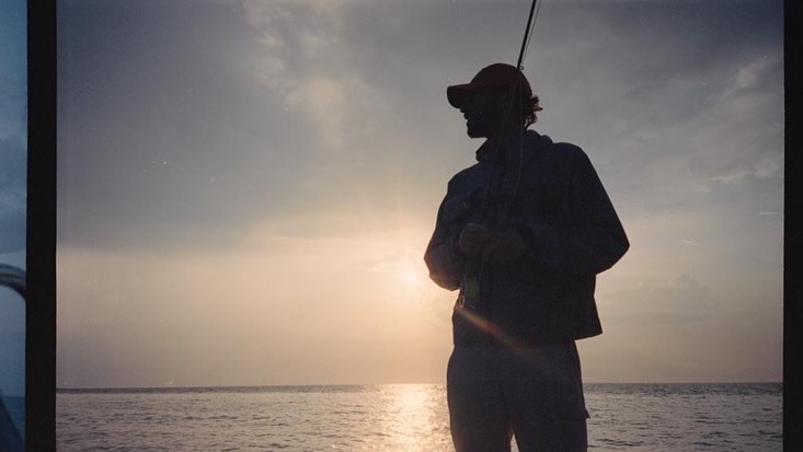 Silhouette of a man wearing a baseball cap holding a fishing rod against a sunset sky over the ocean