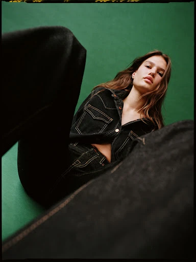 Brunette woman in a dark denim jacket poses against a green background with black denim jeans placed around her in the photo studio.