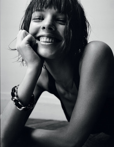 Black and white portrait of a smiling young woman with short hair wearing a chain bracelet and resting her chin on her hand.