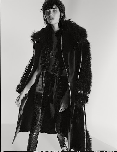 Model in dark faux fur trimmed coat and textured trousers against plain studio backdrop by Matteo Montanari