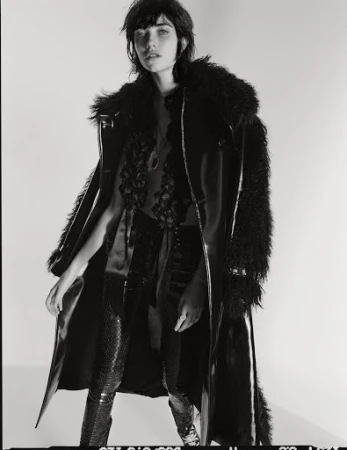 Model in dark faux fur trimmed coat and textured trousers against plain studio backdrop by Matteo Montanari