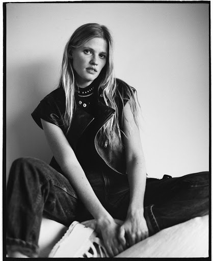 Black and white portrait of a blonde woman in a dark leather vest and jeans seated against a plain wall.