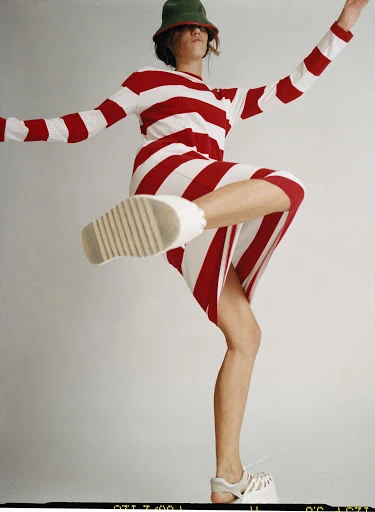 A female fashion model strikes a dynamic pose indoors, wearing a red and white striped long-sleeve dress, green hat, and platform sandals.