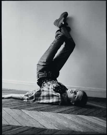 Full-length black and white shot of a child lying upside down on a carpet, resting against a wall with legs straight up in the air.
