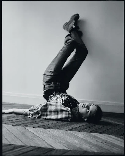 Full-length black and white shot of a child lying upside down on a carpet, resting against a wall with legs straight up in the air.
