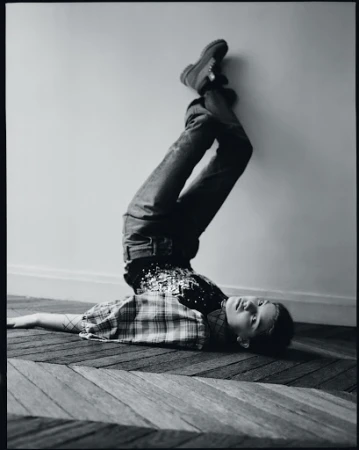 Full-length black and white shot of a child lying upside down on a carpet, resting against a wall with legs straight up in the air.