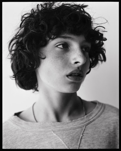 Monochrome portrait of a young adult with curly hair and freckles, wearing a grey sweatshirt, gazing slightly off-center against a light background.