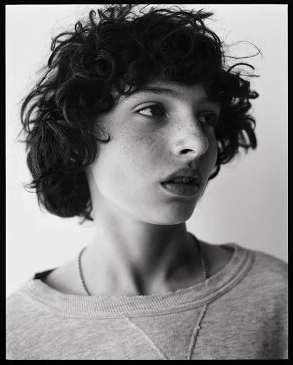 Monochrome portrait of a young adult with curly hair and freckles, wearing a grey sweatshirt, gazing slightly off-center against a light background.
