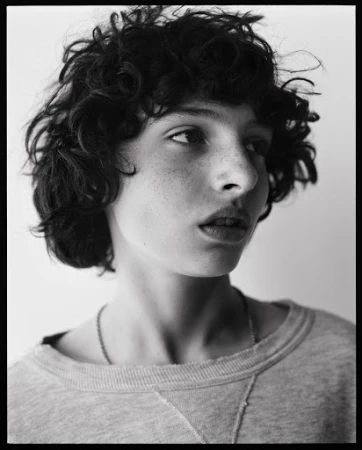 Monochrome portrait of a young adult with curly hair and freckles, wearing a grey sweatshirt, gazing slightly off-center against a light background.