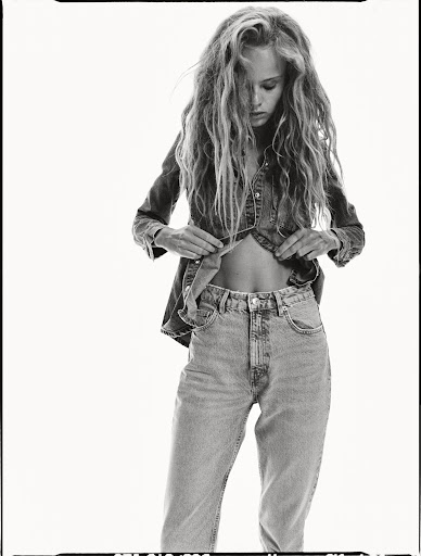Black and white fashion shot of a young adult female model in denim top and jeans, open at the midriff, in a photo studio setting.