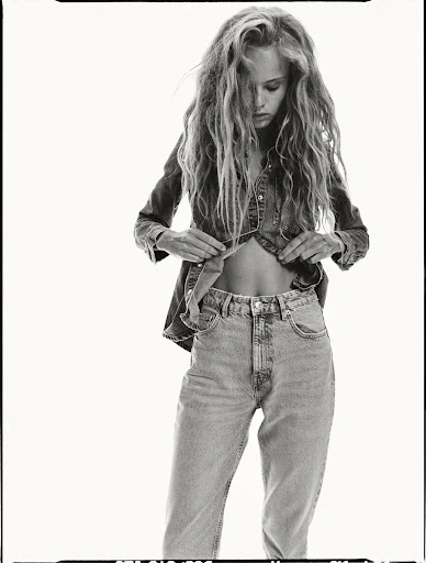 Black and white fashion shot of a young adult female model in denim top and jeans, open at the midriff, in a photo studio setting.