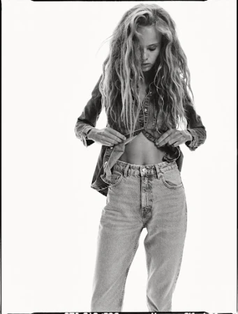 Black and white fashion shot of a young adult female model in denim top and jeans, open at the midriff, in a photo studio setting.