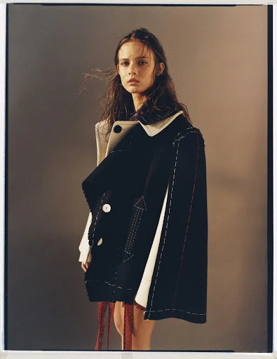 Young woman in deconstructed black and white jacket posing indoors against neutral background in a fashion editorial style image.