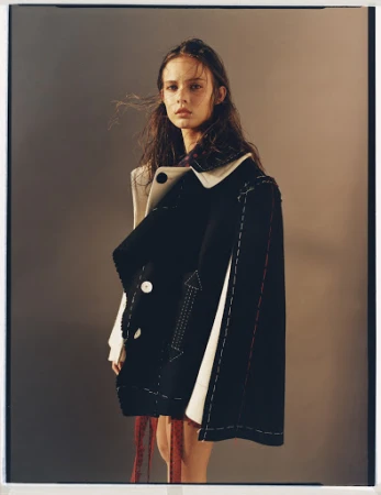 Young woman in deconstructed black and white jacket posing indoors against neutral background in a fashion editorial style image.