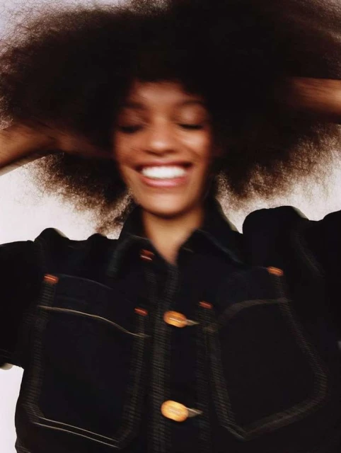 Smiling woman with afro poses in denim jacket in studio, bright lighting, shallow focus portrait, fashion editorial photography.