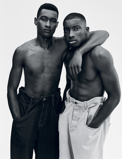 Black and white studio shot of two shirtless adult male fashion models in baggy jeans posing together, one with arm around the other.