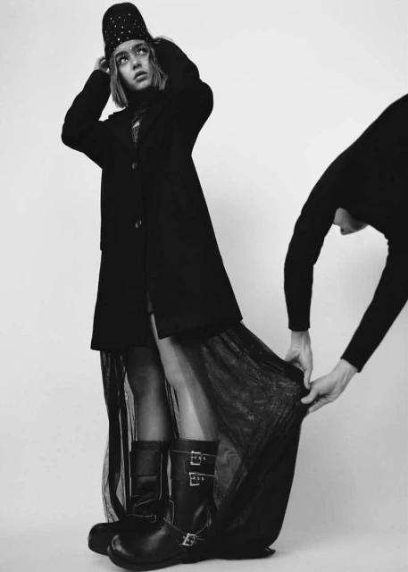Monochrome studio shot of a woman in a hat and coat, assisted by another person adjusting her sheer black skirt. Bold fashion.