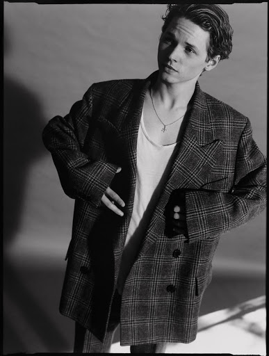 Portrait of a young adult male in an oversized plaid suit jacket and white undershirt, striking a pose in a studio setting.