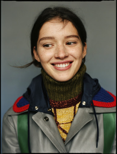 Smiling woman portrait. Close-up of a young adult female with freckles wearing a colorful knit sweater and jacket against a gray background.