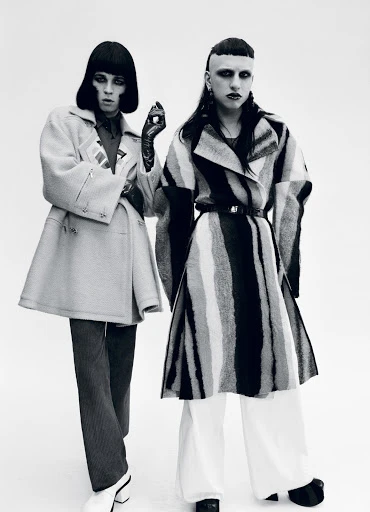 Two fashion models in coats and wide-legged pants pose in a studio. One wears dark tones and black gloves, while the other has white pants.