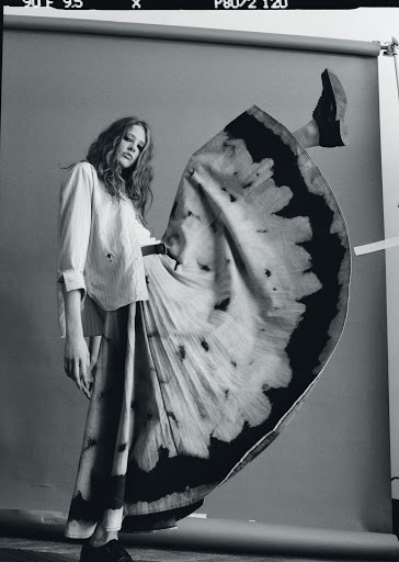 Fashion shot of a woman in a long skirt and collared shirt posing with one leg up in a studio with a gray background.